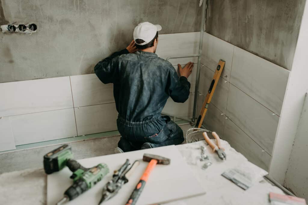 Un ouvrier installe des carreaux muraux blancs sur un mur gris dans une salle de bain en construction, avec outils au premier plan.
