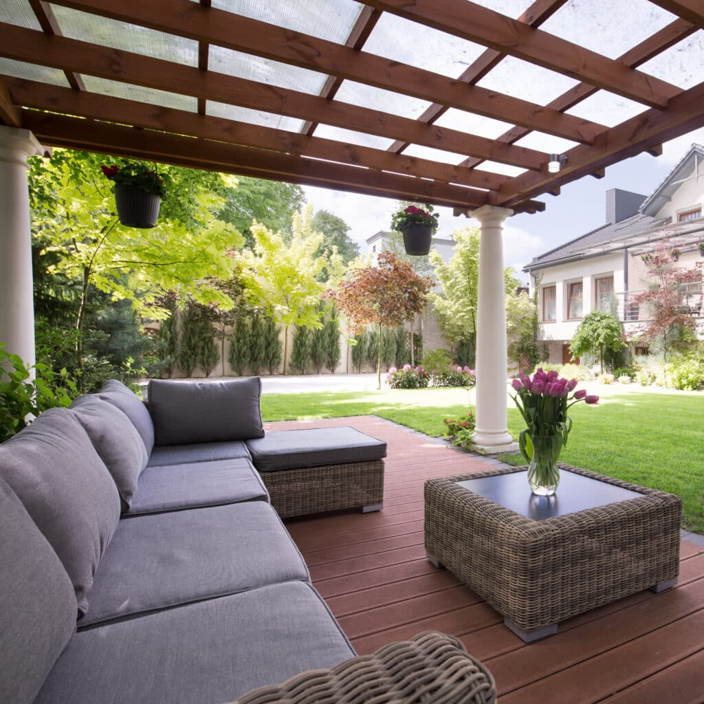 Patio moderne sous pergola bois-transparent. Salon de jardin gris en rotin: canapé d'angle, table basse avec tulipes roses. Grande pelouse et maison.