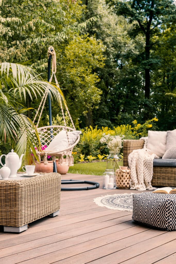 Terrasse en bois avec salon de jardin en osier, fauteuil suspendu, théière blanche et pouf motif. Ambiance cozy dans un jardin verdoyant.